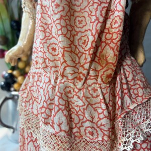 May include: A doll wearing a dress made of light-colored fabric with a repeating orange floral pattern. The dress features a gathered waistline, a ruffled skirt section, and delicate cream-colored lace trim along the bottom hem of the garment.