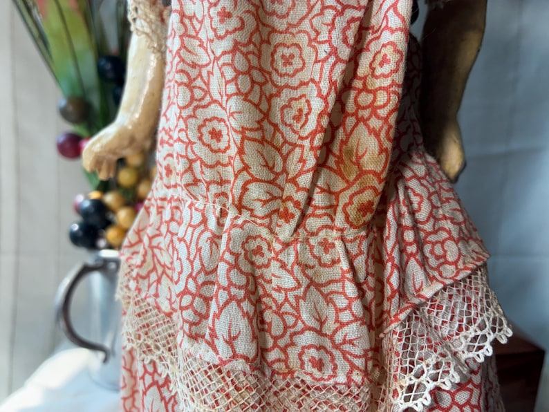 May include: A doll wearing a dress made of light-colored fabric with a repeating orange floral pattern. The dress features a gathered waistline, a ruffled skirt section, and delicate cream-colored lace trim along the bottom hem of the garment.