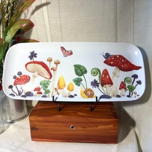 May include: White rectangular serving tray with a whimsical mushroom design. The tray features red, yellow, and brown mushrooms, a snail, and a butterfly. The tray is supported by a black metal stand.