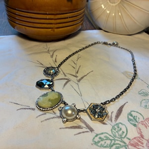 May include: A silver-toned chain necklace featuring five distinct charms: a small round crystal, a dark faceted hexagon, a large oval cameo with a light green profile, a round white faux pearl, and a hexagonal floral-shaped metal charm.