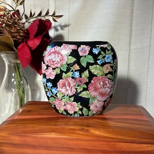 May include: A rounded ceramic vase with a glossy black background featuring a colorful floral pattern of pink peonies, green leaves, and small blue flowers. The vase has a wide, open top and a smooth, curved body that tapers slightly toward the base.