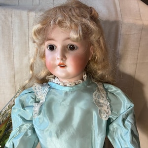 Op de afbeelding: Een vintage porseleinen pop met blond haar en roze wangen. De pop draagt een lichtblauwe satijnen jurk met kanten rand. De ogen van de pop zijn donker en expressief, en de lippen zijn lichtjes geopend.
