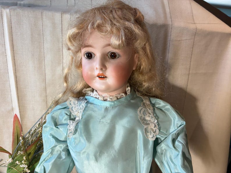 Op de afbeelding: Een vintage porseleinen pop met blond haar en roze wangen. De pop draagt een lichtblauwe satijnen jurk met kanten rand. De ogen van de pop zijn donker en expressief, en de lippen zijn lichtjes geopend.