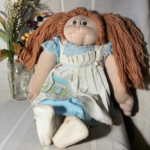 May include: A vintage doll with a soft, round face, green eyes, and long, reddish-brown yarn hair styled in pigtails. The doll wears a light blue dress with a white apron and white shoes. The doll is sitting on a light-colored surface.