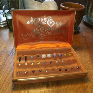 May include: Open orange jewelry box with gold trim, filled with various earrings. The box has a floral design on the inside lid. The earrings include studs, hoops, and dangle styles in various colors and shapes.