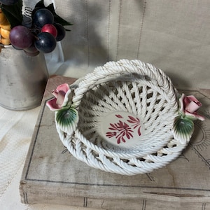 May include: A white ceramic basket with a woven design and a handle. The basket is decorated with pink roses and green leaves. The interior features a floral design in red. A metal pitcher with faux grapes is in the background.