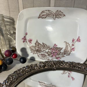 May include: White rectangular ceramic plate with a floral design in brown and pink. The plate has rounded corners and a slightly raised edge. The floral pattern includes leaves and small flowers. A decorative silver-toned tray is in the foreground.