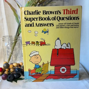 May include: A vintage hardcover book titled "Charlie Brown's Third Super Book of Questions and Answers." The cover is yellow with illustrations of Charlie Brown, Snoopy, a train, and an airplane. The book's theme is about transportation.