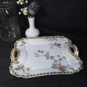 May include: A white porcelain serving tray with gold trim and floral accents. The rectangular tray features handles and a scalloped edge. A clear glass jar and a small white vase with flowers are in the background.
