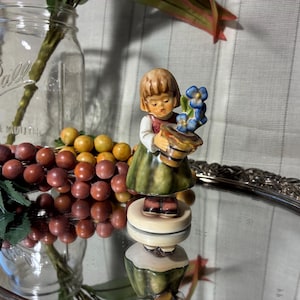 May include: A vintage figurine of a young girl holding a small pot of blue flowers. The girl wears a green dress, red vest, and has blonde hair. The figurine is on a decorative stand, with a reflective surface.
