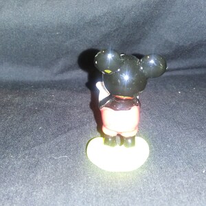 May include: A small Mickey Mouse figurine, viewed from the back, with a black head and ears, red shorts, and yellow shoes. The figure is standing on a yellow base. The background is a dark, textured surface.