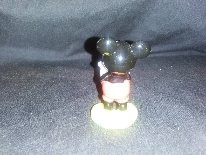 May include: A small Mickey Mouse figurine, viewed from the back, with a black head and ears, red shorts, and yellow shoes. The figure is standing on a yellow base. The background is a dark, textured surface.