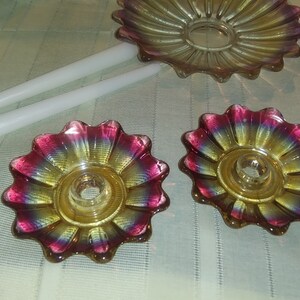 May include: Two iridescent glass candle holders shaped like flowers with a yellow centre and pink and purple edges. The candle holders are on a white surface.