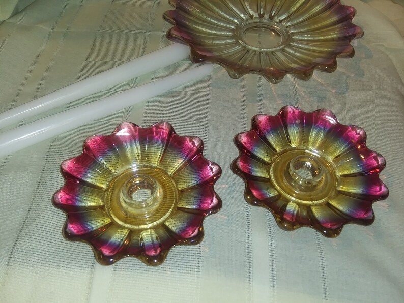 May include: Two iridescent glass candle holders shaped like flowers with a yellow centre and pink and purple edges. The candle holders are on a white surface.