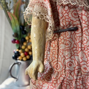 May include: A doll with a textured, light-colored arm and hand, wearing a garment made of light-colored fabric with a repeating orange floral pattern. The dress features intricate lace trim on the sleeves and hem, and a metal pin is visible.