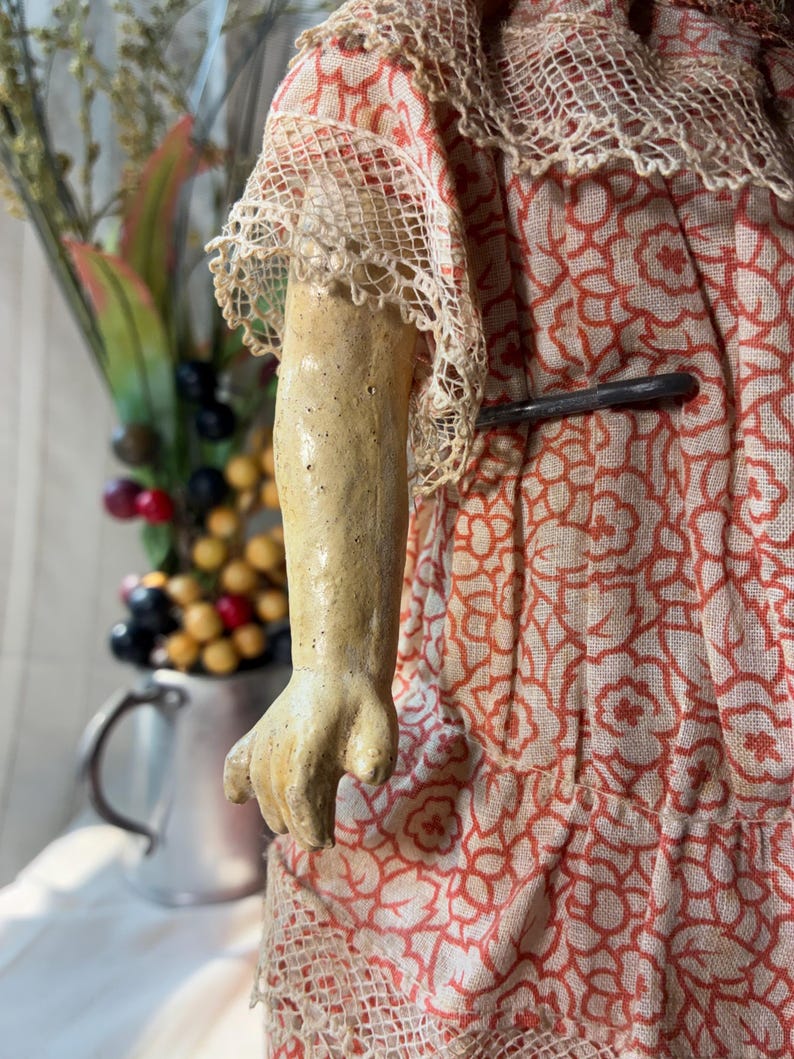May include: A doll with a textured, light-colored arm and hand, wearing a garment made of light-colored fabric with a repeating orange floral pattern. The dress features intricate lace trim on the sleeves and hem, and a metal pin is visible.