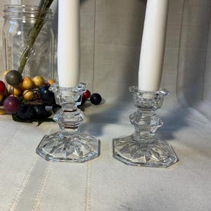 May include: Two clear crystal candle holders with white candles. The candle holders have a faceted design and a hexagonal base. The background includes a glass jar with berries and greenery.