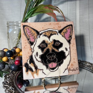 May include: Square ceramic tile featuring a German Shepherd dog portrait. The dog has a black nose, brown eyes, and pink-lined ears. The tile has a speckled, light brown background and a white border. The dog's face is centered on the tile.
