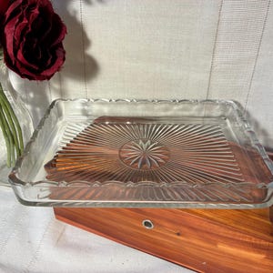 May include: A rectangular clear glass tray featuring a pressed sunburst pattern radiating from the center and a scalloped edge. The tray is positioned above a wooden box, with a clear glass vase containing deep red artificial flowers nearby.