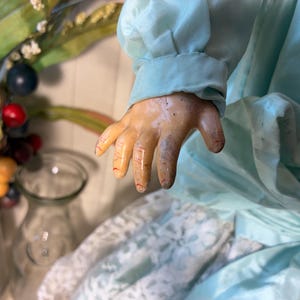 Op de afbeelding: Een vintage pop in een lichtblauwe jurk, met een gedetailleerde hand. De hand van de pop toont geschilderde vingers en een verouderd uiterlijk. De jurk heeft een satijnachtige textuur. Een glazen vaas en decoratieve bessen staan op de achtergrond.