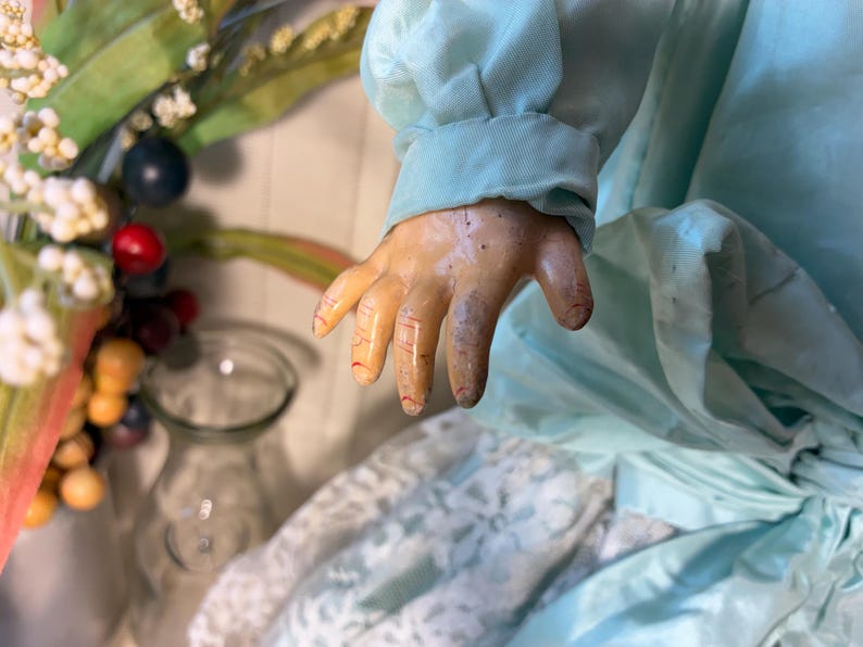 Op de afbeelding: Een vintage pop in een lichtblauwe jurk, met een gedetailleerde hand. De hand van de pop toont geschilderde vingers en een verouderd uiterlijk. De jurk heeft een satijnachtige textuur. Een glazen vaas en decoratieve bessen staan op de achtergrond.