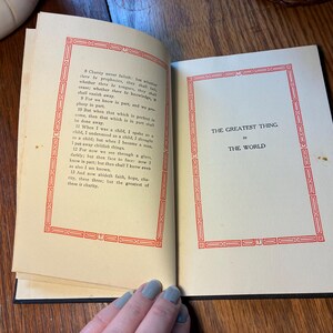 May include: An open book with cream-coloured, aged pages. The left page has text within a red decorative border. The right page displays the title "THE GREATEST THING IN THE WORLD" within a similar red border. The book is on a wooden surface.