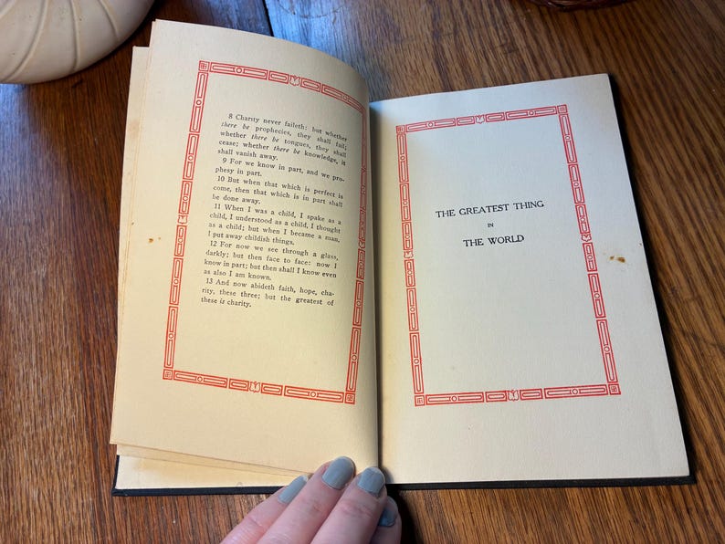 May include: An open book with cream-coloured, aged pages. The left page has text within a red decorative border. The right page displays the title "THE GREATEST THING IN THE WORLD" within a similar red border. The book is on a wooden surface.