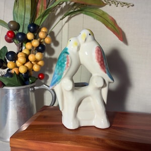 May include: A ceramic figurine featuring two white birds perched together on a branch. The bird on the left has blue and red wing accents, while the bird on the right has red wing accents. Both birds have yellow heads and black eyes.