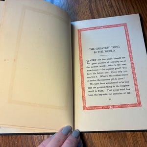 May include: An open book with aged, yellowed pages. The right page features text within a red decorative border, with the title "THE GREATEST THING IN THE WORLD." The text is in black, with a serif font. The left page is blank.
