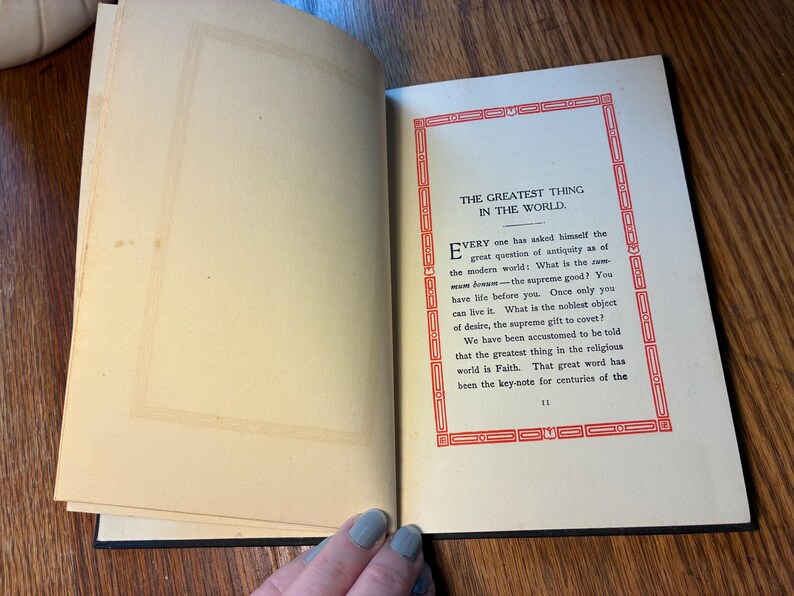 May include: An open book with aged, yellowed pages. The right page features text within a red decorative border, with the title "THE GREATEST THING IN THE WORLD." The text is in black, with a serif font. The left page is blank.