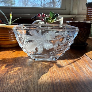 May include: A clear, square-shaped glass bowl with etched floral designs. The bowl sits on a wooden surface, with other decorative pots and plants in the background. The bowl's edges are slightly textured.