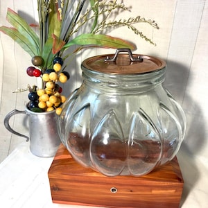 May include: A clear glass jar with a wide, rounded body and vertical fluted panels. The jar features a flat, circular copper-toned lid with a small metal handle centered on top. The glass is transparent, showcasing a smooth, reflective surface.