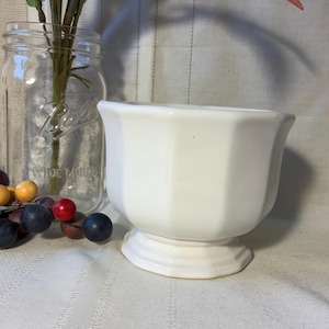 May include: A white ceramic mug with a black handle and the text "But First, Coffee" in a bold, black font. The mug is set against a blurred background, suggesting a cozy setting.