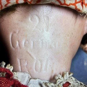 May include: A close-up of the back of a doll's neck featuring the embossed text 21 Germany. The doll has light skin and is wearing a fabric collar with an orange geometric pattern, a white lace trim, and thin red ribbon accents tied around the neck.