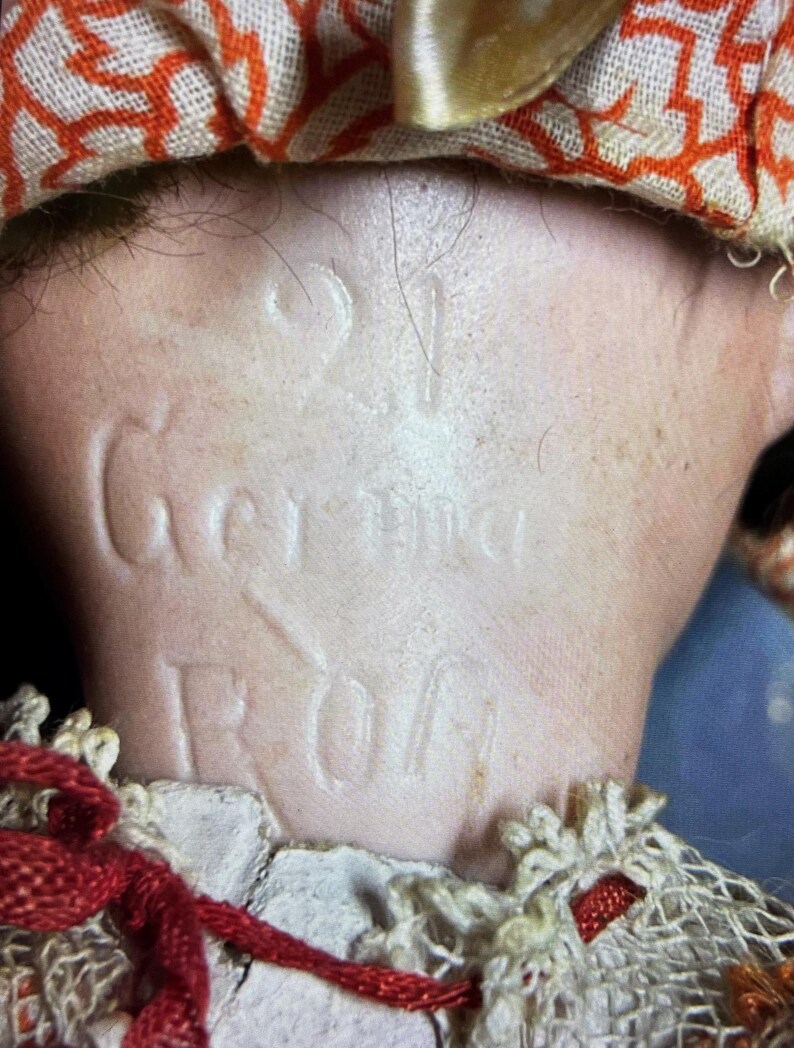 May include: A close-up of the back of a doll's neck featuring the embossed text 21 Germany. The doll has light skin and is wearing a fabric collar with an orange geometric pattern, a white lace trim, and thin red ribbon accents tied around the neck.