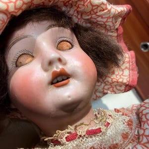 May include: A doll with a pale face, rosy cheeks, and brown hair, wearing a fabric bonnet and dress featuring a red and white floral pattern. The doll has closed eyes with painted lashes, an open mouth showing teeth, and a cream-colored lace collar.