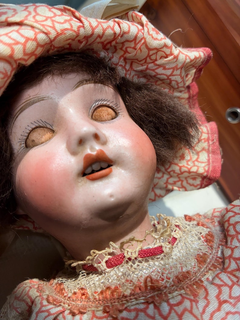 May include: A doll with a pale face, rosy cheeks, and brown hair, wearing a fabric bonnet and dress featuring a red and white floral pattern. The doll has closed eyes with painted lashes, an open mouth showing teeth, and a cream-colored lace collar.