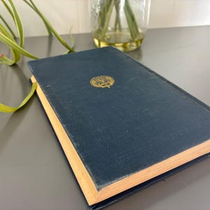 May include: A navy blue hardcover book with gold lettering and a decorative emblem. The book's pages have a golden hue, and the cover shows signs of wear. The book is resting on a gray surface.