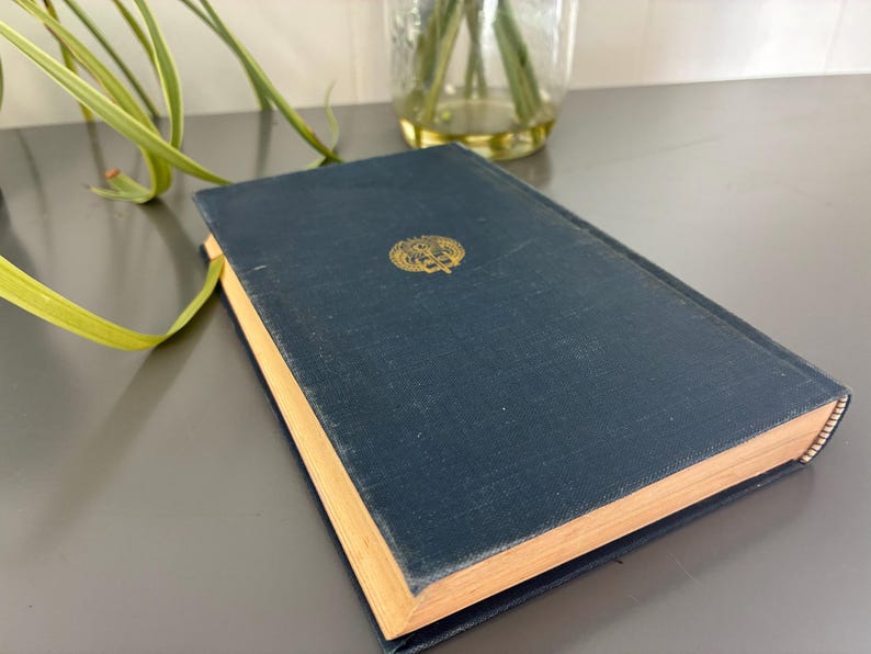 May include: A navy blue hardcover book with gold lettering and a decorative emblem. The book's pages have a golden hue, and the cover shows signs of wear. The book is resting on a gray surface.