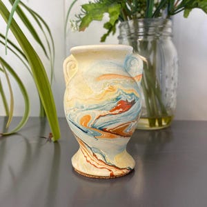 May include: A small, decorative vase with a marbled design in shades of yellow, orange, blue, and white. The vase has a rounded body, a flared base, and two small handles. The vase is approximately 4 inches tall.