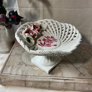 May include: A white ceramic basket with a woven design, resting on a pedestal base. The basket is adorned with pink and green floral accents and a decorative ceramic disc with a red floral pattern. The basket is placed on top of an antique book.
