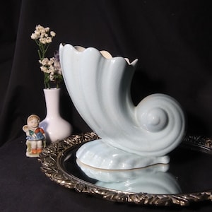 May include: A light blue ceramic vase shaped like a seashell. The vase is sitting on a silver tray with a mirrored surface.