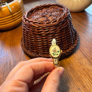 May include: A gold-toned, vintage bracelet watch with an oval face featuring a floral design in pink, green, and white. The watch has a hinged bangle style and is held against a wooden surface. A woven basket is in the background.