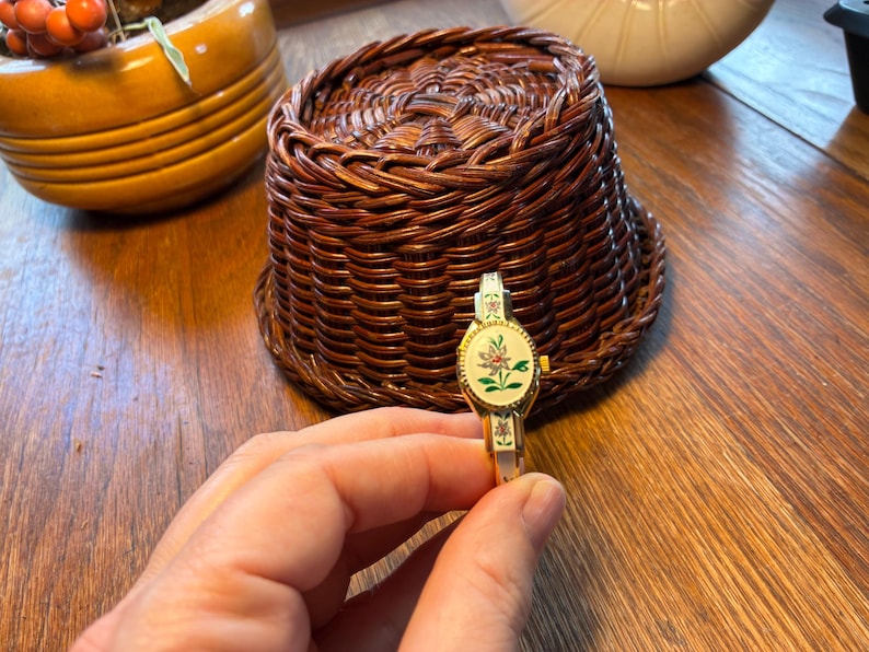 May include: A gold-toned, vintage bracelet watch with an oval face featuring a floral design in pink, green, and white. The watch has a hinged bangle style and is held against a wooden surface. A woven basket is in the background.