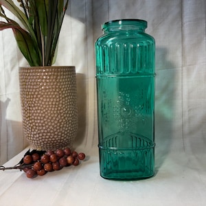 May include: A tall, teal-colored glass jar with a rectangular base and a decorative grape design. The jar is next to a textured brown vase with greenery and a sprig of faux grapes. The jar is approximately 12 inches tall.