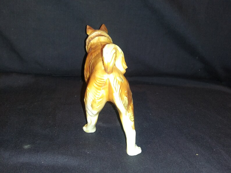 May include: A ceramic figurine of a brown and white dog standing on all four legs. The dog is facing away from the camera and has its tail curled up.