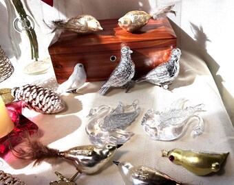 May include: A collection of glass bird and swan ornaments featuring metallic silver, gold, and clear finishes. Several birds have attached feathers and metal clips. Two pinecone-shaped ornaments are also present on a white fabric surface.