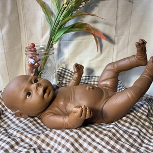 May include: A brown-skinned baby doll lying on a brown and white checkered blanket. The doll has its arms and legs raised. A glass jar with red berries and greenery is in the background.