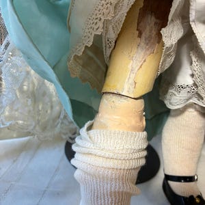 Op de afbeelding: Close-up van de benen van een vintage pop. De pop heeft een geschilderd been met afbladderende verf, draagt witte gebreide sokken en zwarte lakleren schoenen met een bandje en een kleine strik.