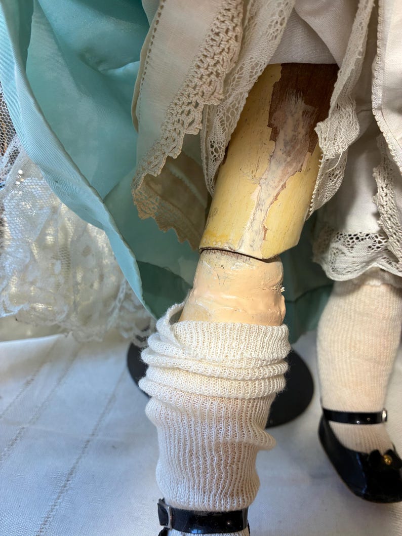 Op de afbeelding: Close-up van de benen van een vintage pop. De pop heeft een geschilderd been met afbladderende verf, draagt witte gebreide sokken en zwarte lakleren schoenen met een bandje en een kleine strik.
